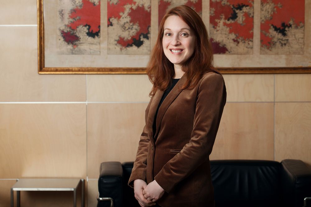 Professional portrait in brown blazer with decorative artwork visible in background.