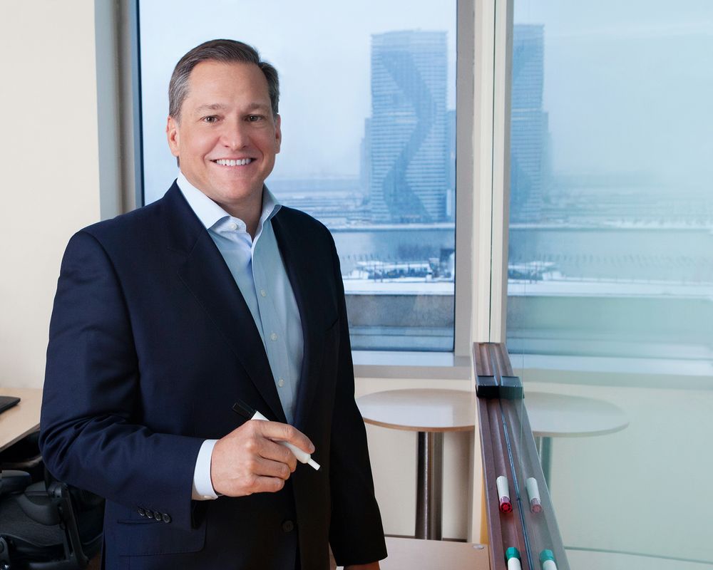 Professional portrait of person in dark suit standing by office window with city view.