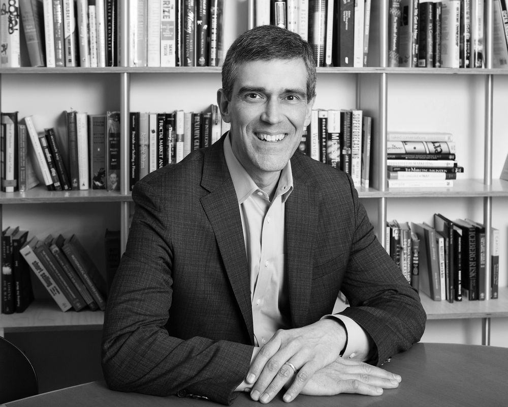 Professional headshot of person in dark suit sitting at desk with bookshelf in background.