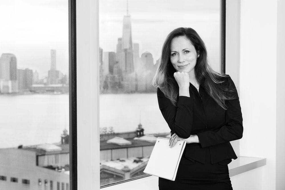 Black and white portrait of person in business attire standing by window with city skyline view.