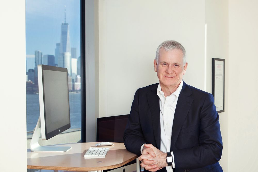 Professional office portrait with city skyline and computer monitor visible in background.