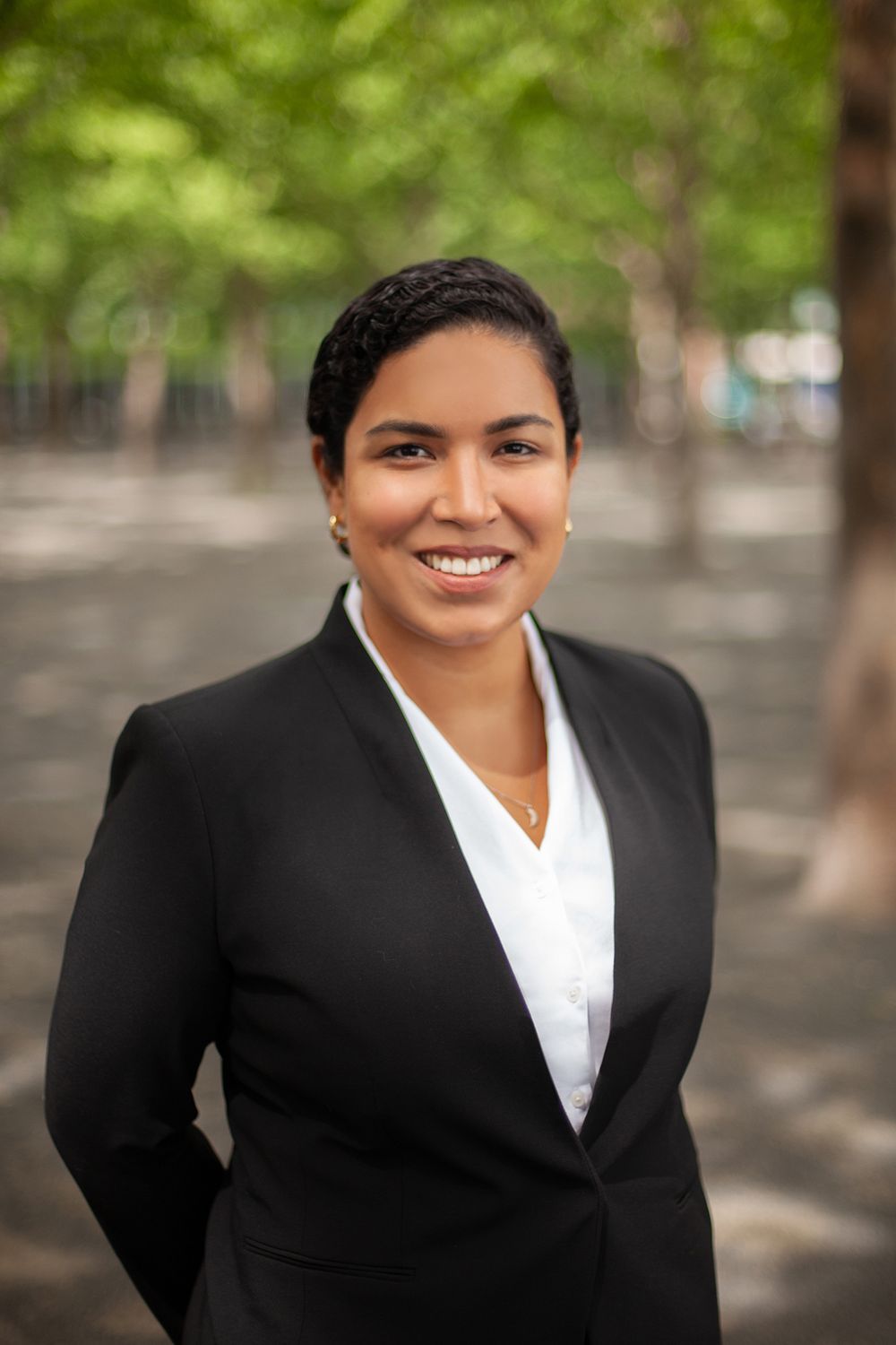 Professional portrait of person in black business suit and white blouse against blurred outdoor background.
