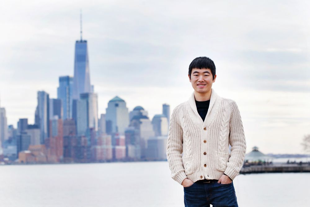 Person in cream cardigan standing with New York City skyline and One World Trade Center in background.