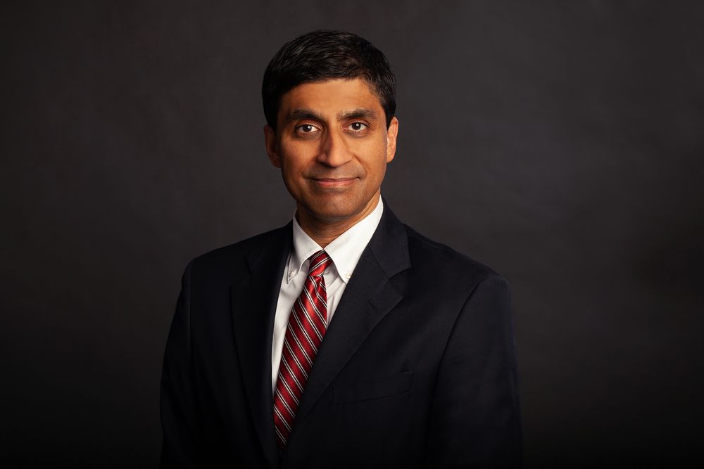 Professional headshot portrait in dark suit with red tie against black background.