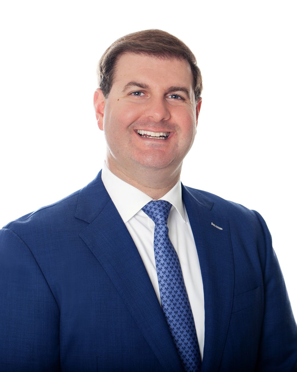 Professional headshot of person wearing navy suit and blue polka dot tie against light background.