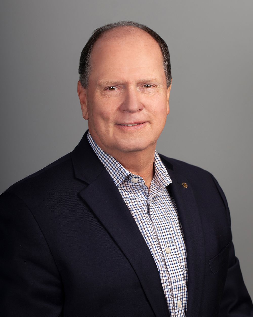 Professional portrait of person in checkered shirt and navy blazer against grey background.