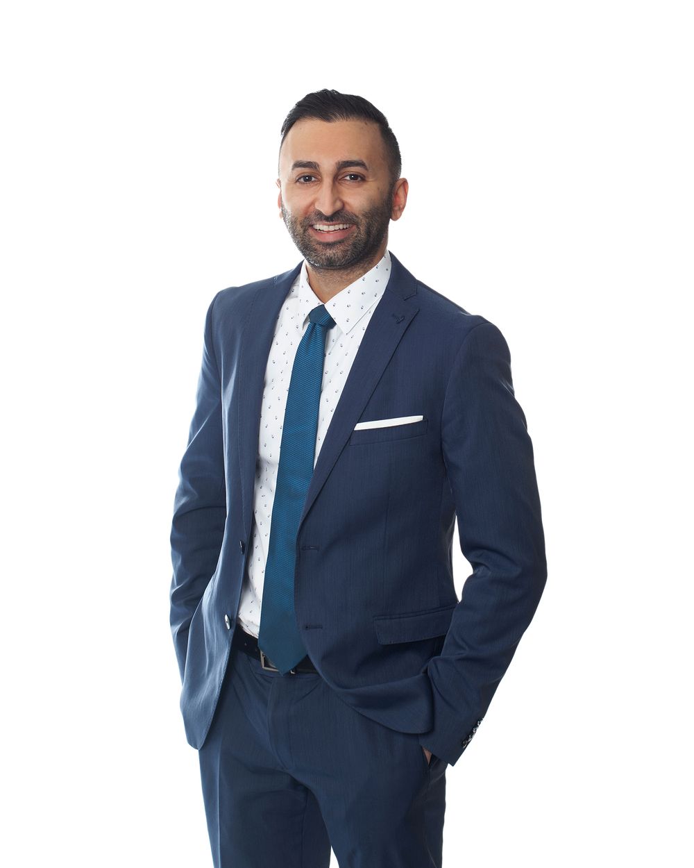 Professional portrait of person in navy suit with hands in pockets against white background.
