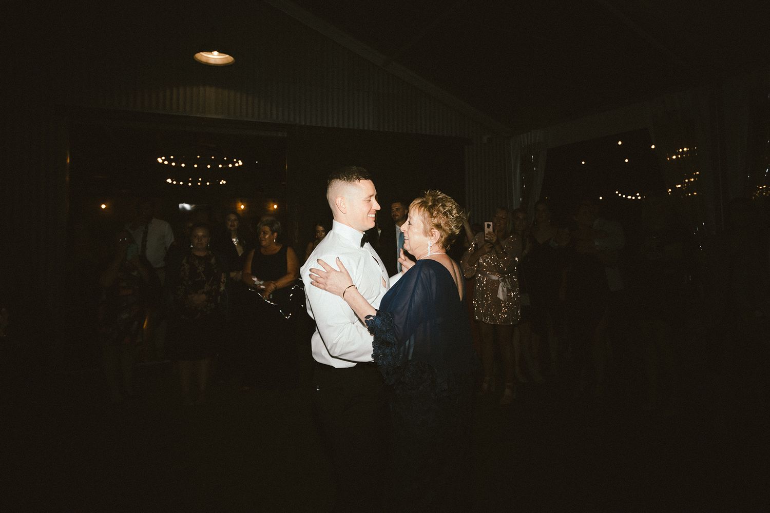 Wedding guests dancing together under decorative string lights at an evening reception.