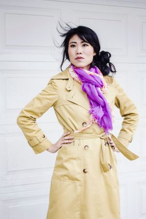 Fashionable model in beige trench coat and purple scarf poses against white wall.
