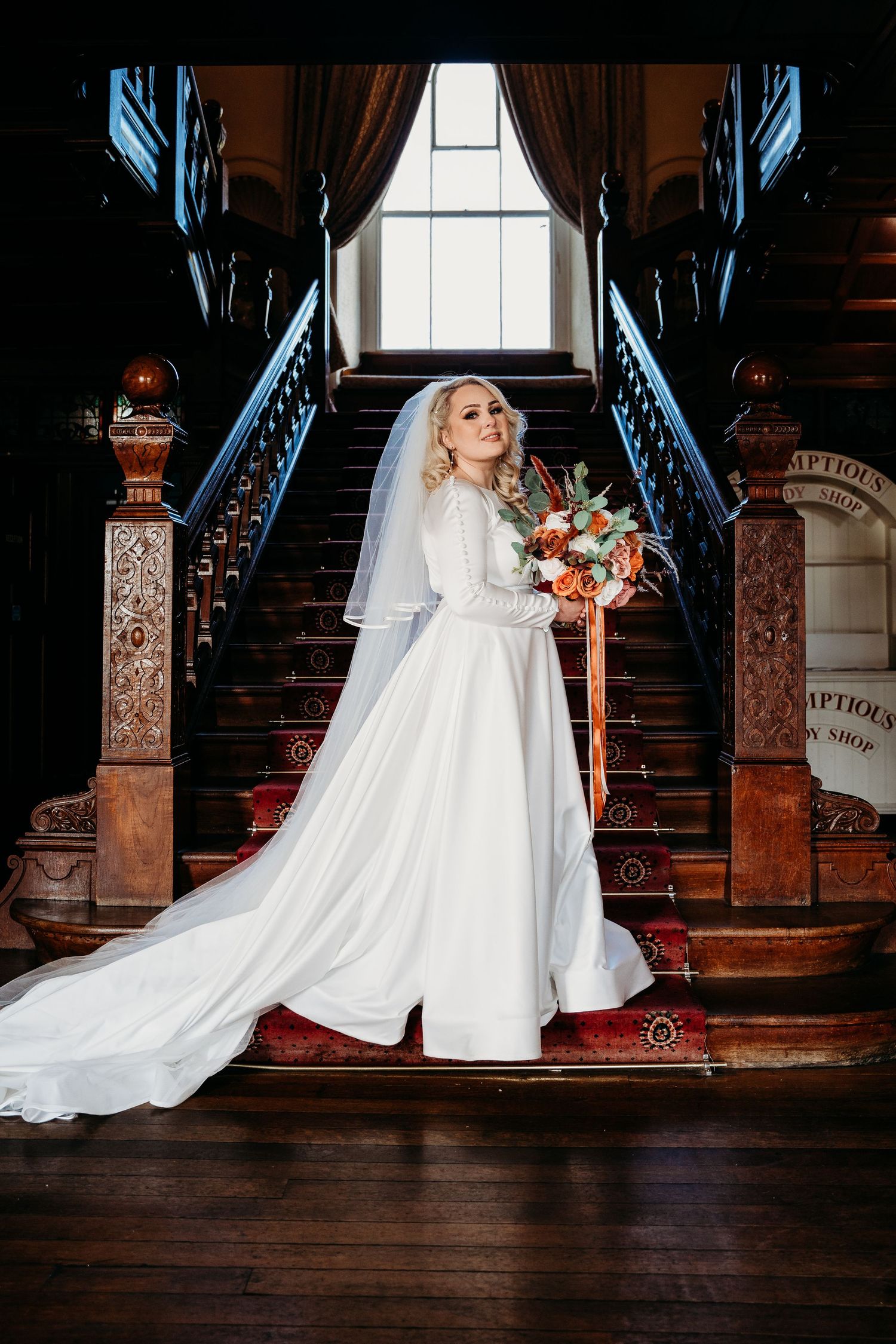 Elegant bridal portrait on vintage staircase with flowing white gown and autumn bouquet.