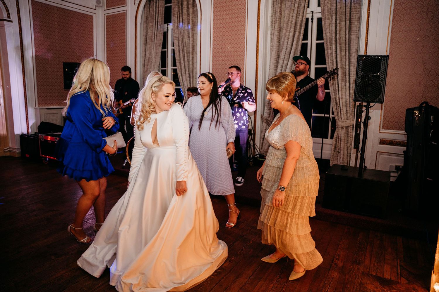 Wedding guests dance and celebrate on a wooden dance floor in an ornate ballroom.