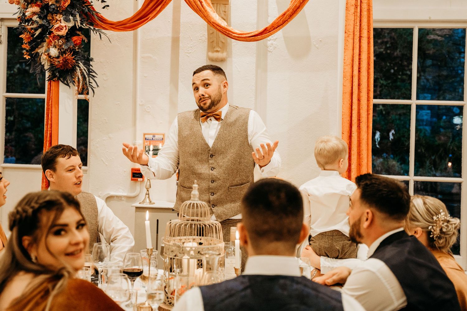Wedding reception speech moment with guests gathered around decorated tables.