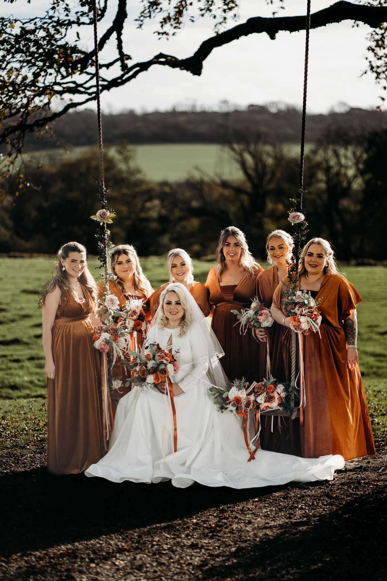 A bridal party in autumn-colored dresses poses outdoors on a rustic swing.