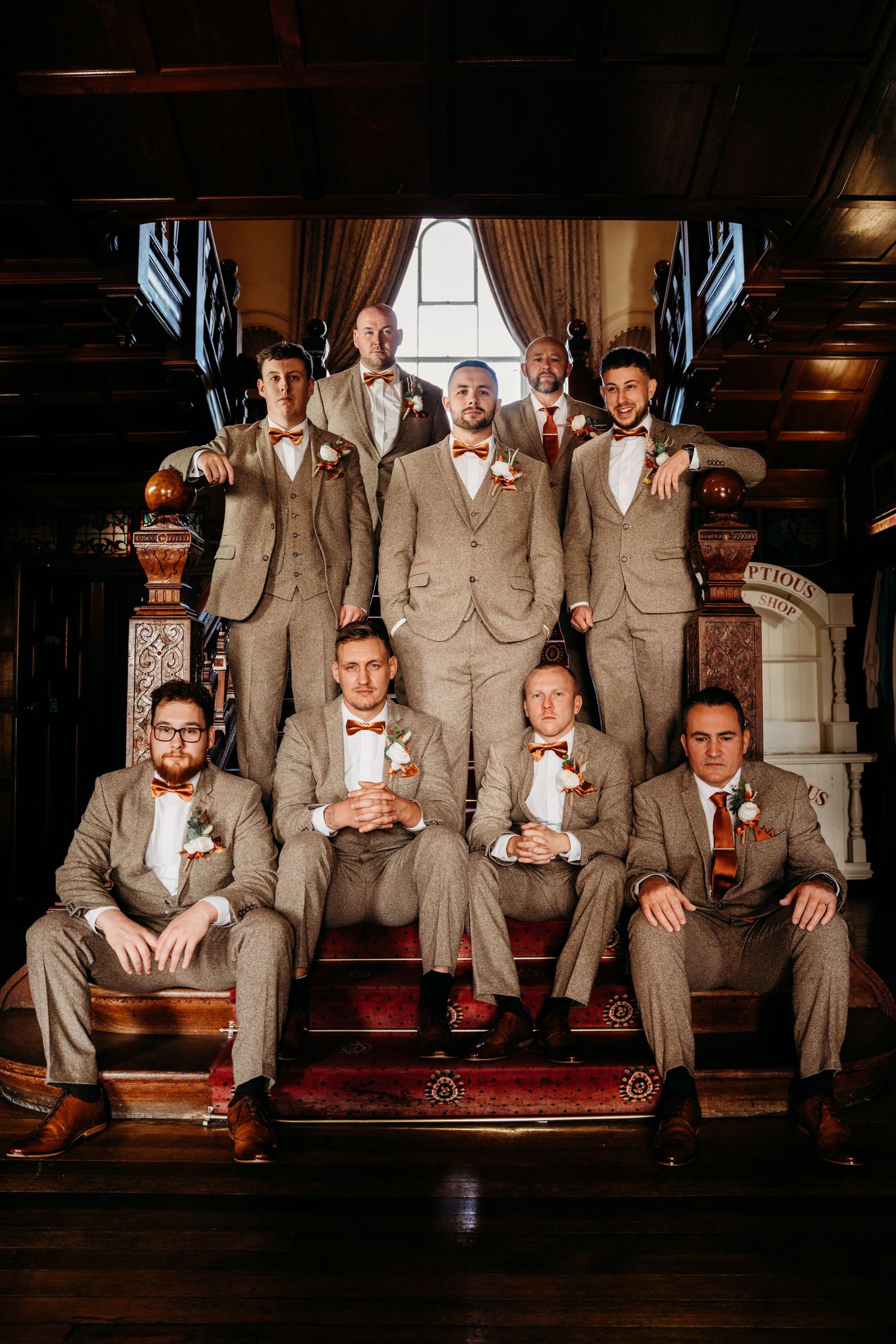Groomsmen in matching tan suits pose confidently on wooden stairs in a dimly lit venue.