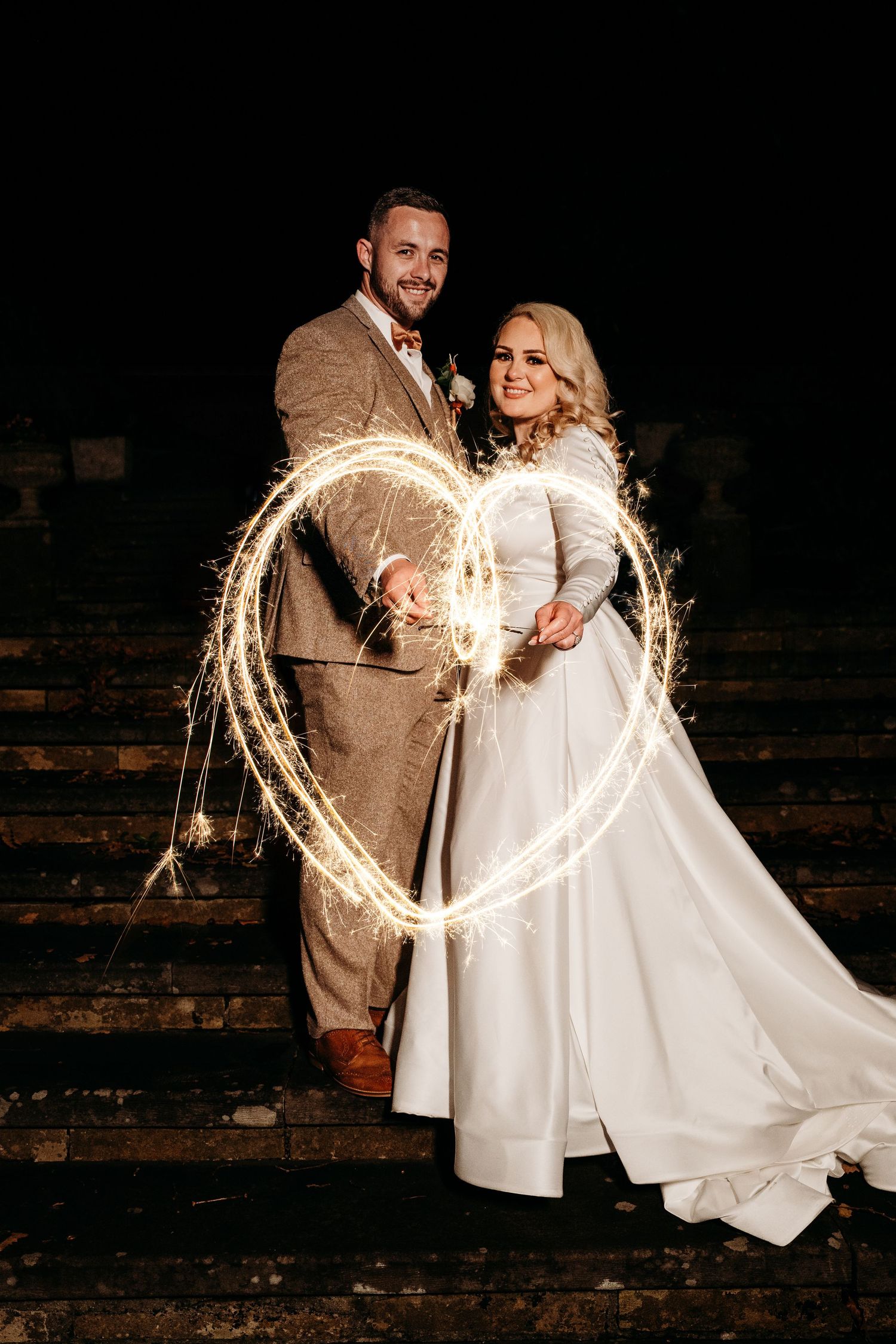 Creative sparkler light painting creates heart shape around couple on wedding night.