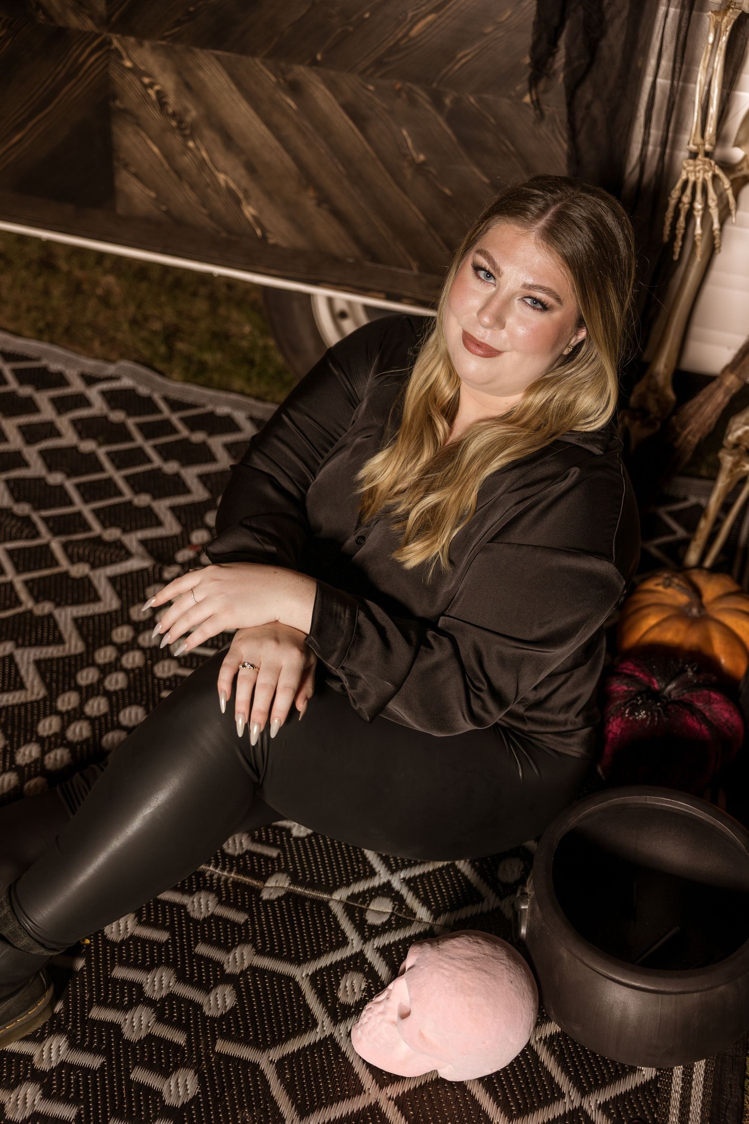 A person in a black outfit sits on a patterned rug with decorative items in a moody indoor setting.