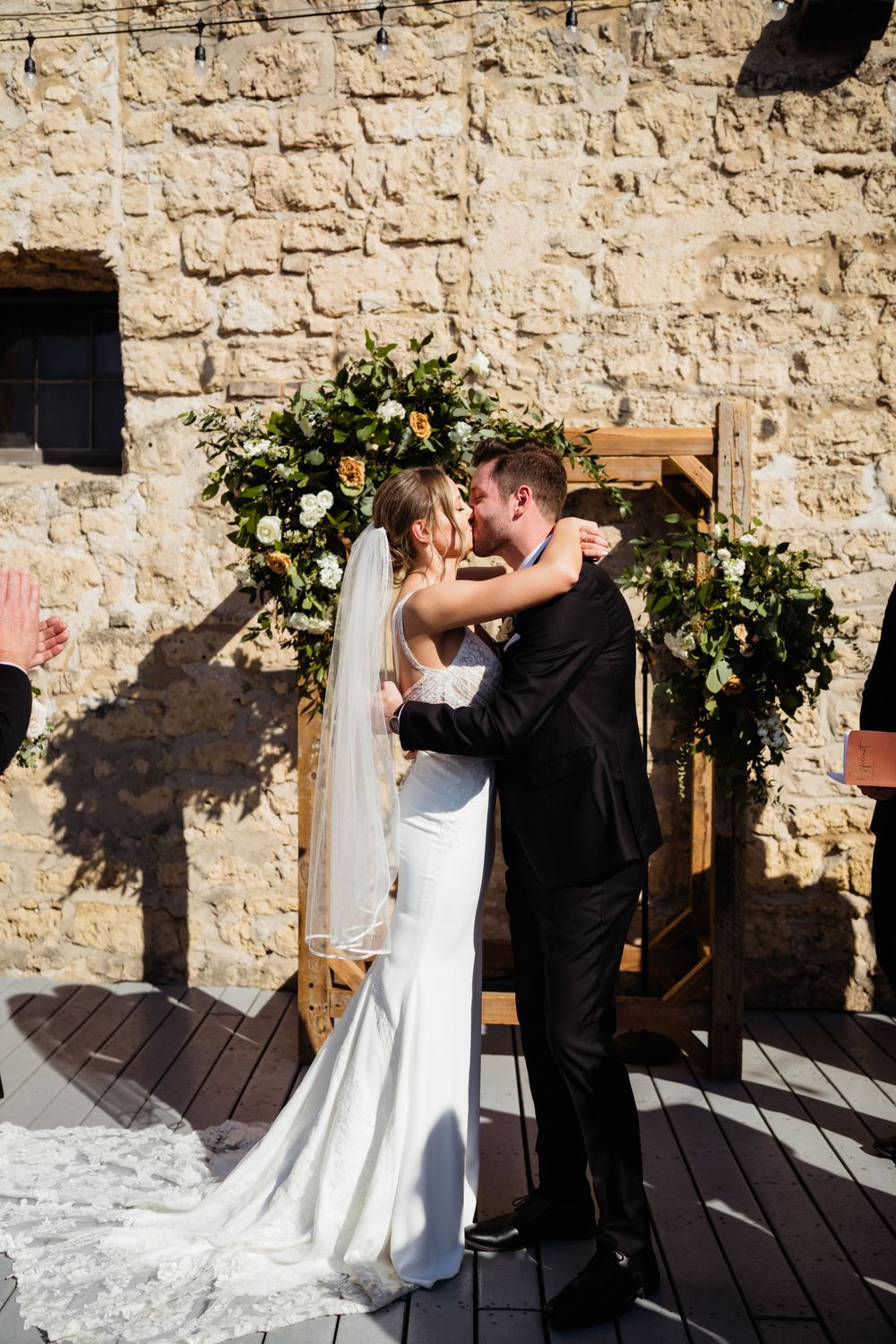 Wedding couple shares intimate moment under floral arch against stone wall.