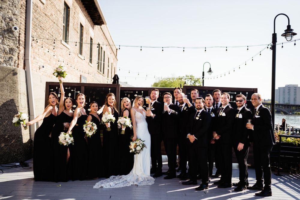 Wedding party in black attire poses outside historic building with string lights.
