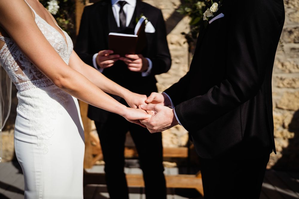 Close up of hands holding during an outdoor wedding ceremony with officiant standing in background.