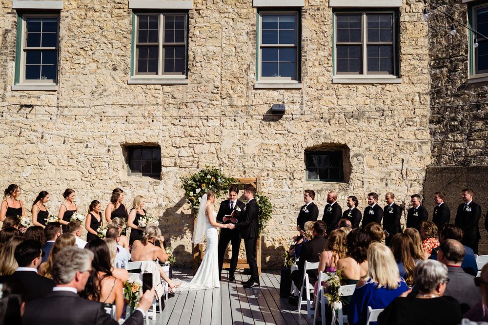 Wedding ceremony taking place against rustic stone wall backdrop with floral arch.