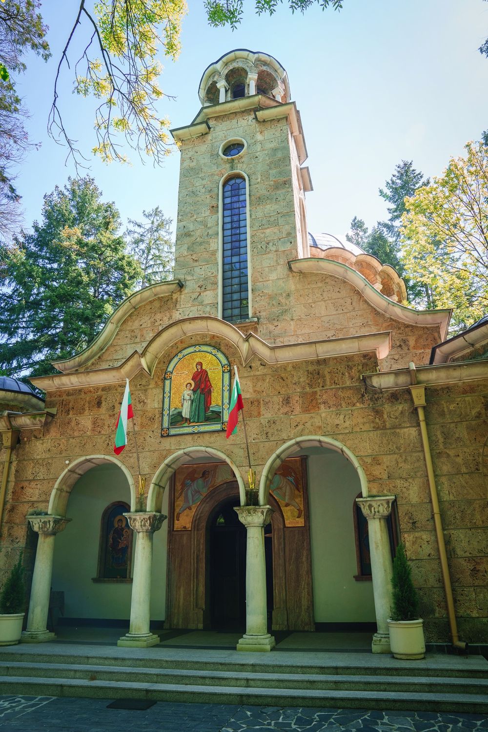 Traditional Orthodox church exterior with stone walls, arched entrance, and bell tower against blue sky.
