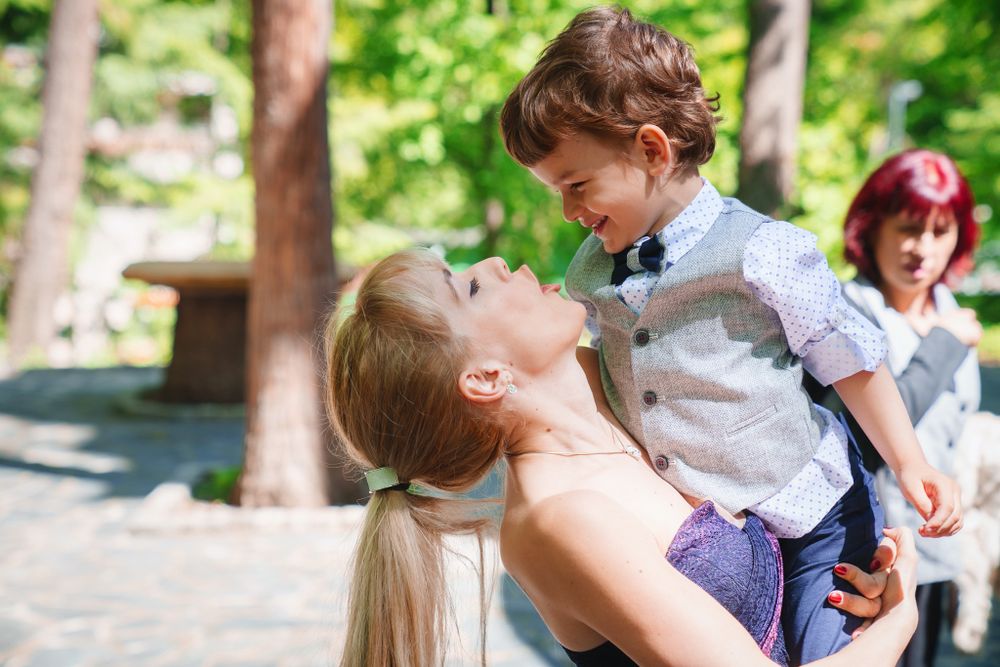 Tender moment captured outdoors with green trees and natural lighting in background.