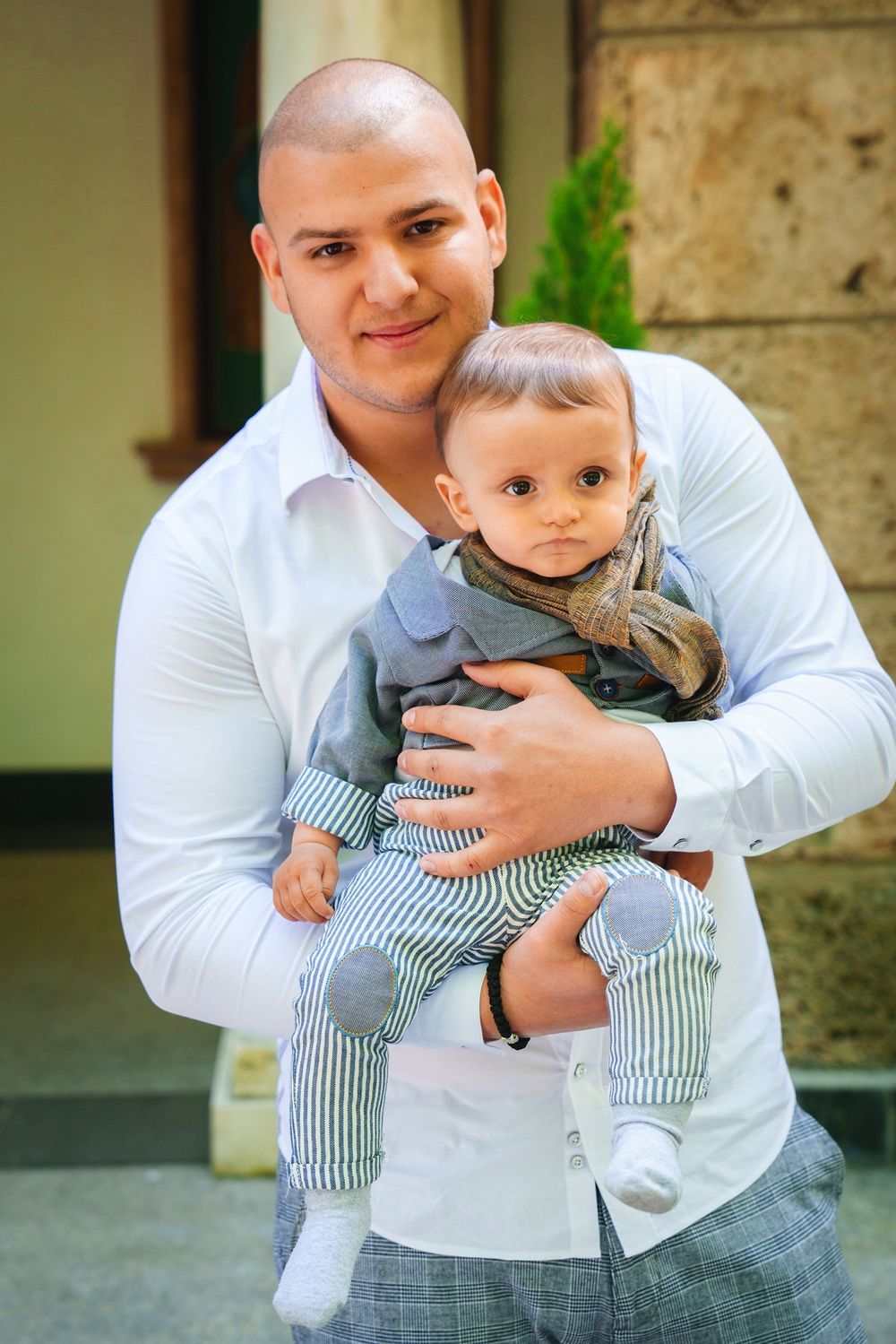 Adult in white shirt holds small baby wearing striped outfit against stone building exterior.