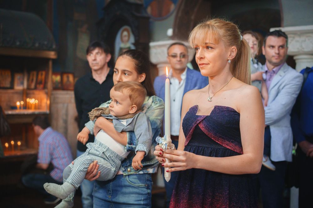 Guests watching a christening ceremony in an Orthodox church with candlelight.