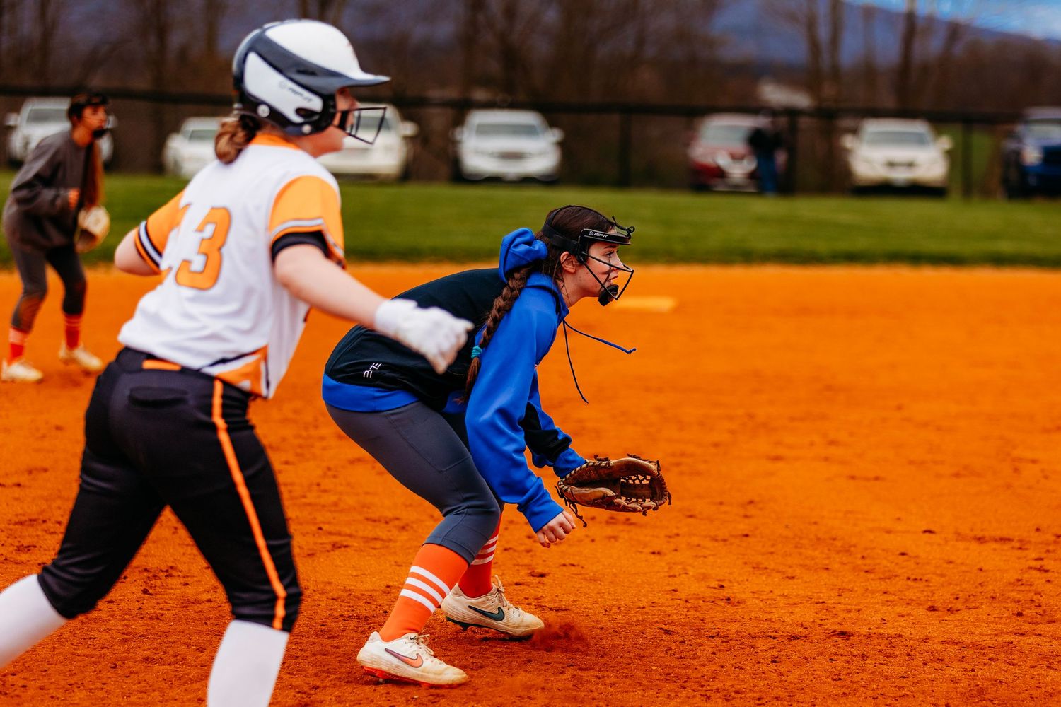 Youth softball player in orange uniform rounds third base during an outdoor game on red clay field.