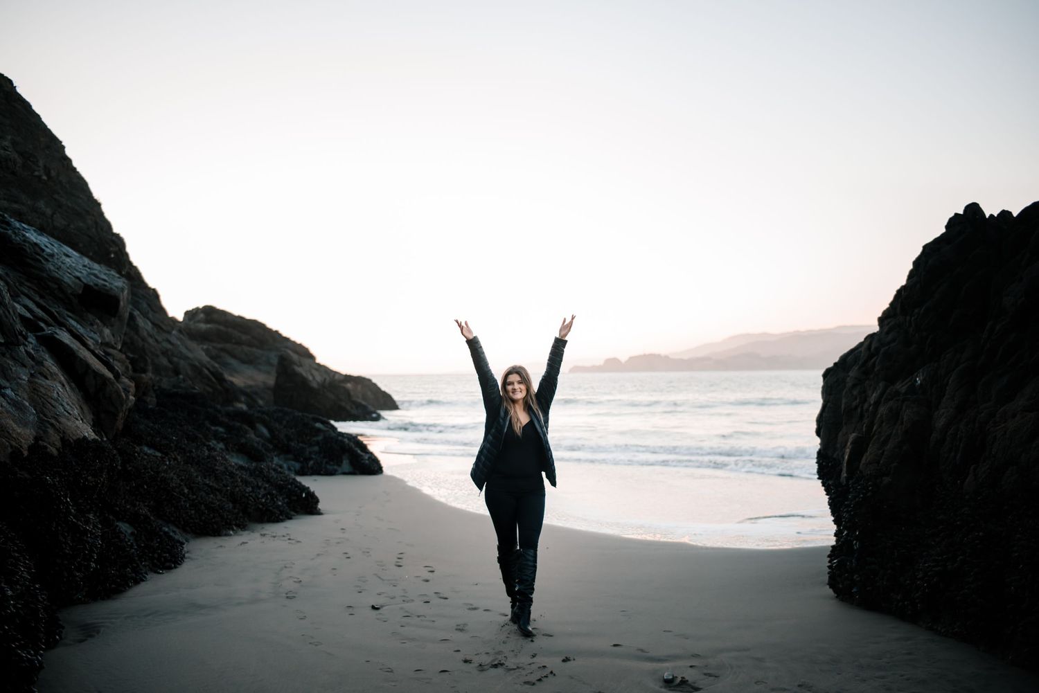 Silhouetted figure stands between rocky cliffs on a misty beach at sunset.