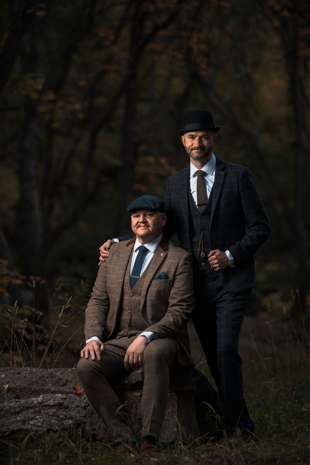 Two people in vintage-inspired suits pose dramatically in moody forest setting.