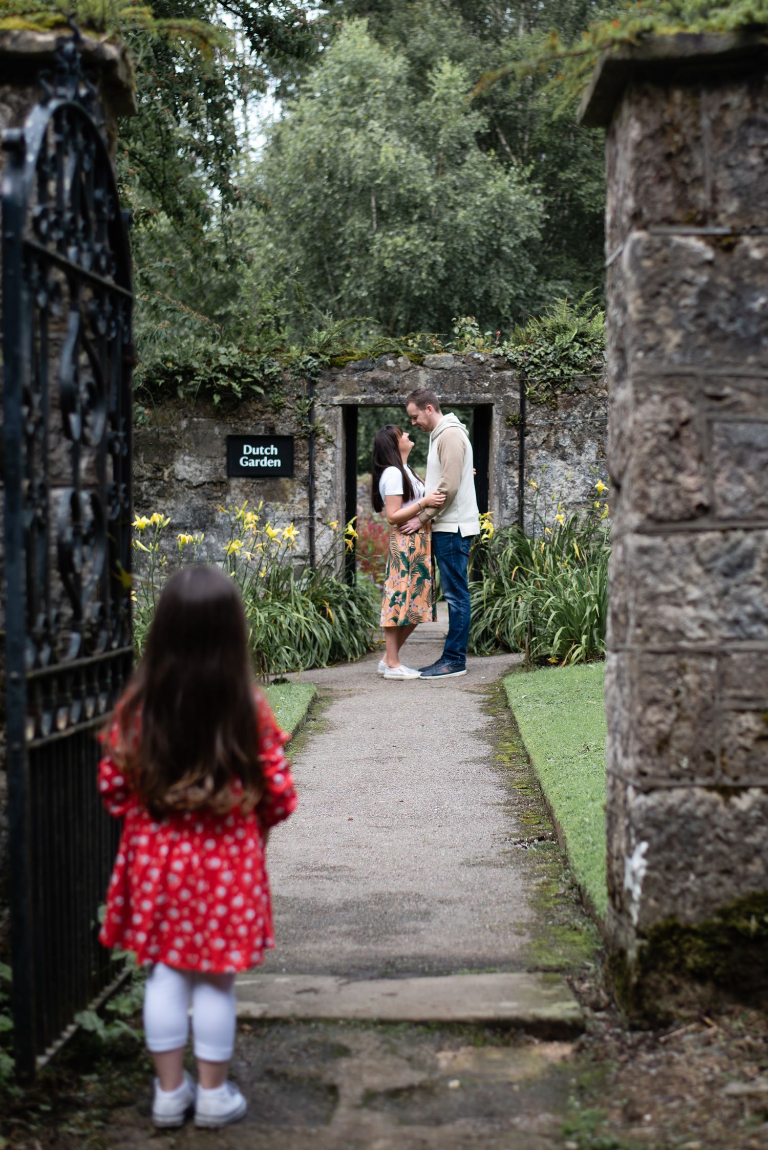 A small child in a red dress watches a couple through an ornate garden gate on a stone pathway.
