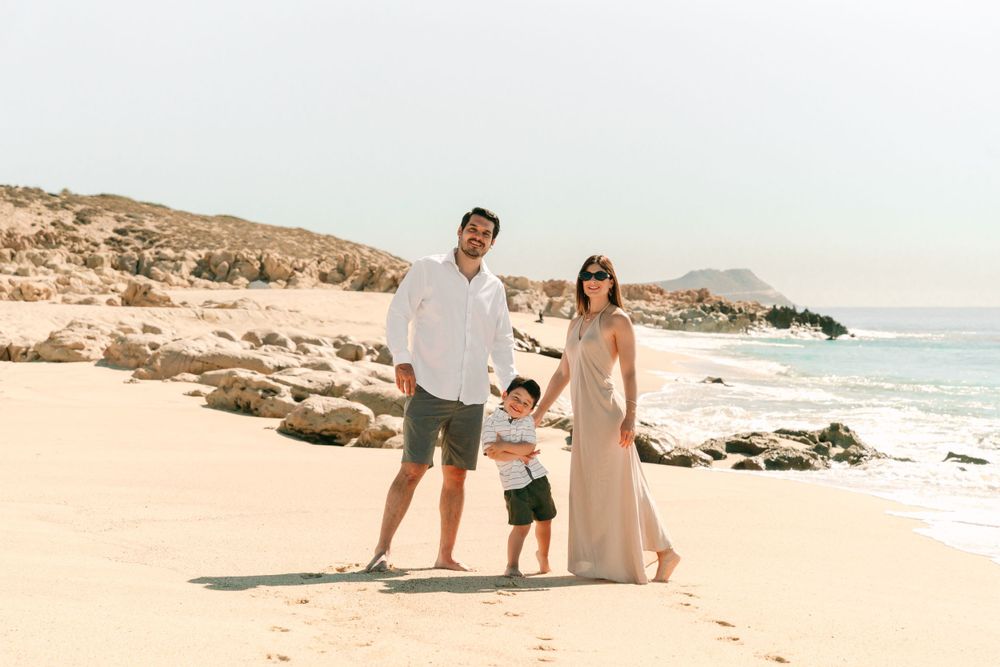 Family of three posing together on scenic beach with rocky coastline.
