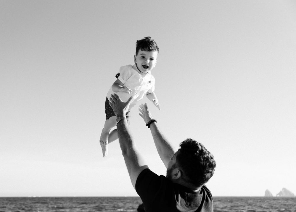 A black and white capture of a playful moment between parent and child against a bright sky.