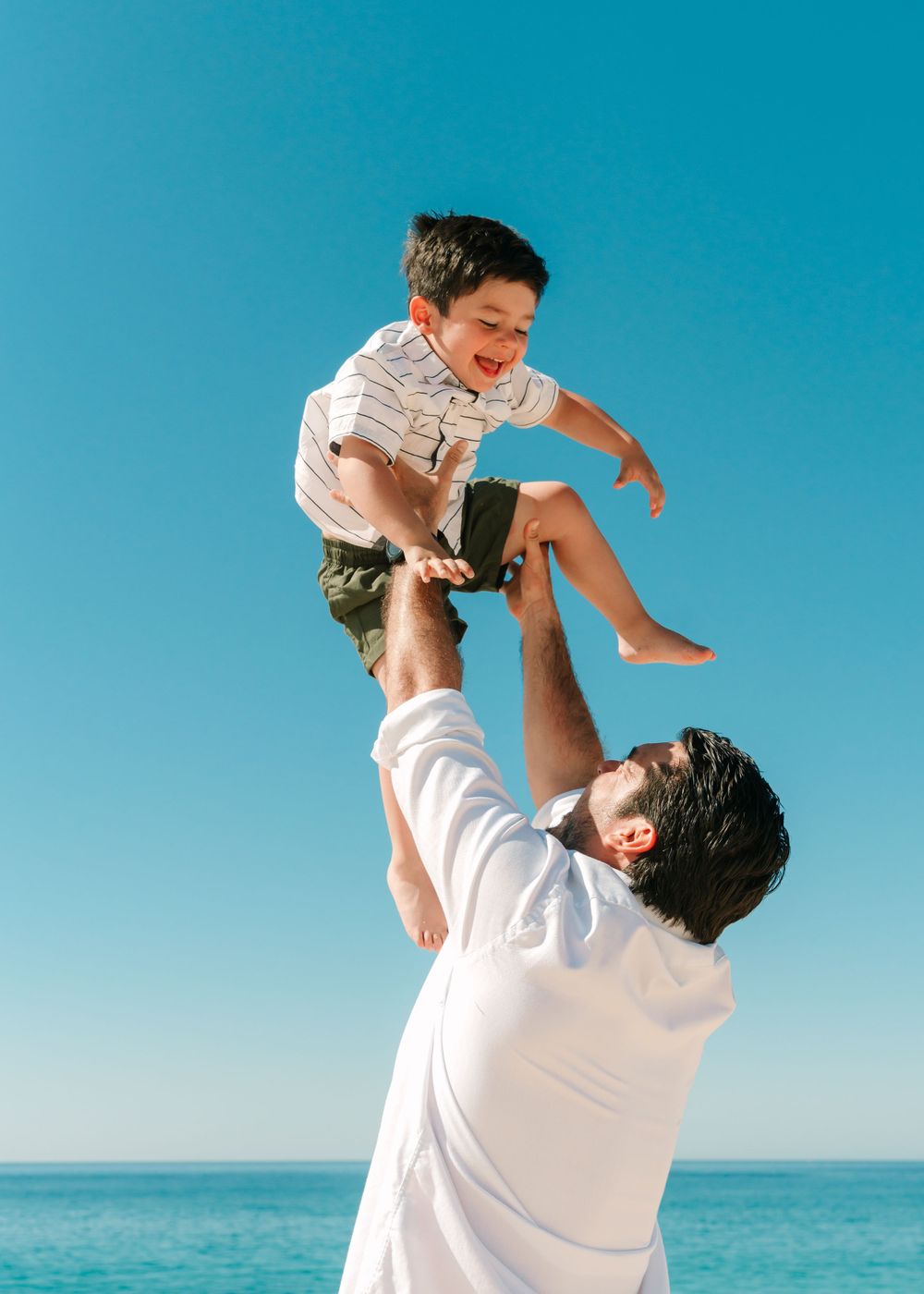 Father lifting child playfully against bright blue sky and ocean backdrop.