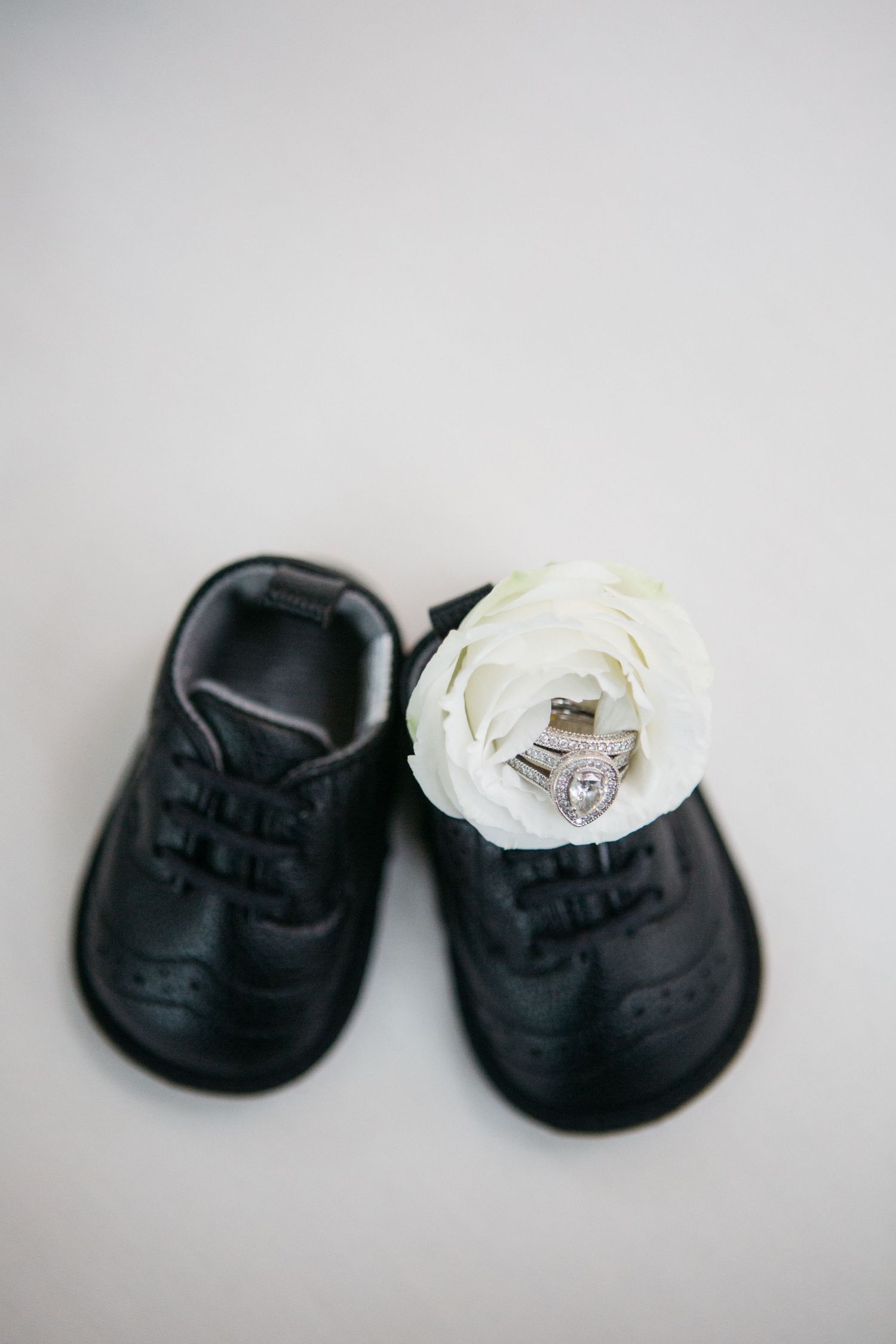 a flat lay of black baby shoes against a white backdrop showcasing a flower and wedding ring