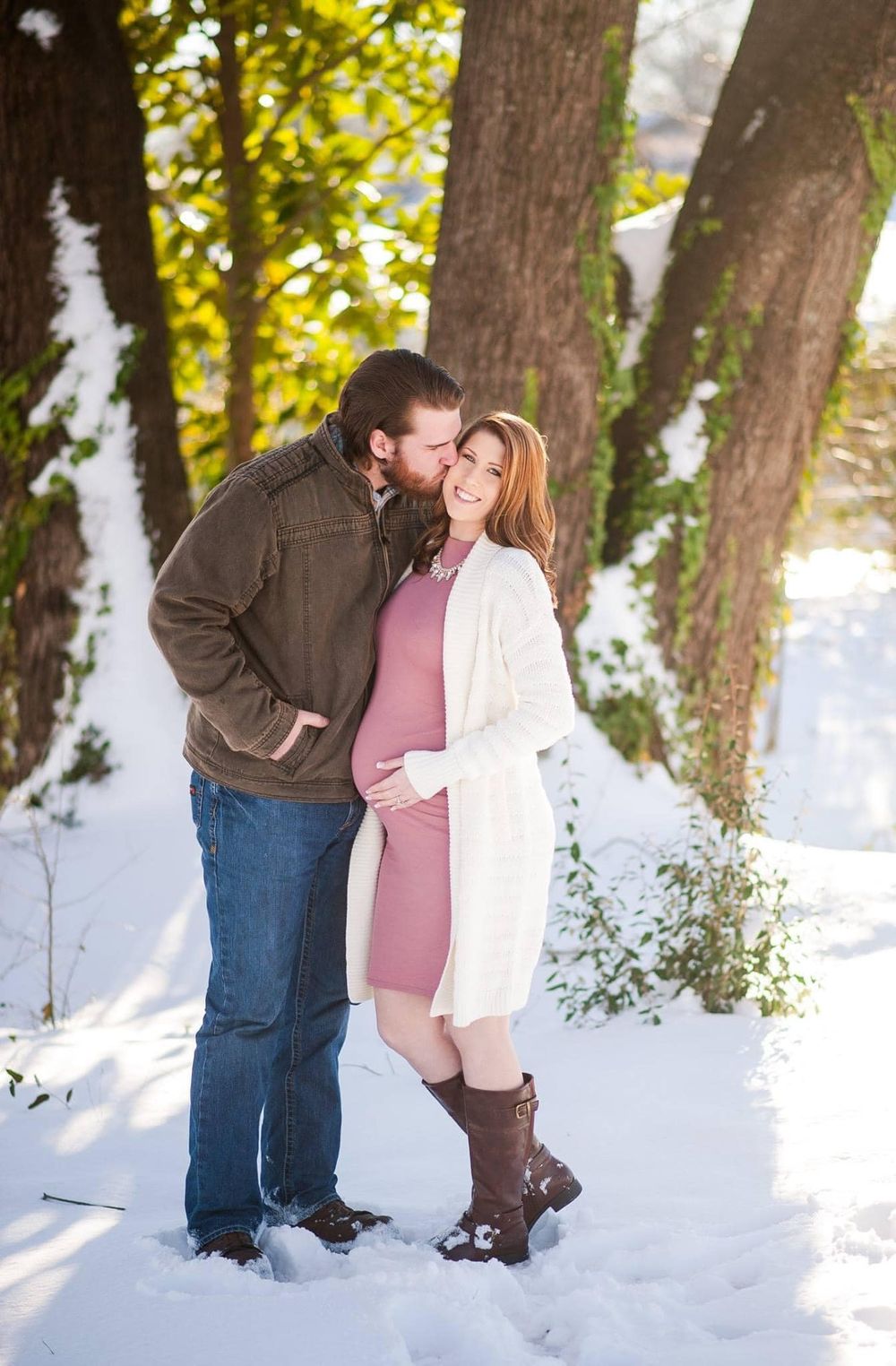 Two people embrace in a snowy wooded area with sunlight filtering through the trees.