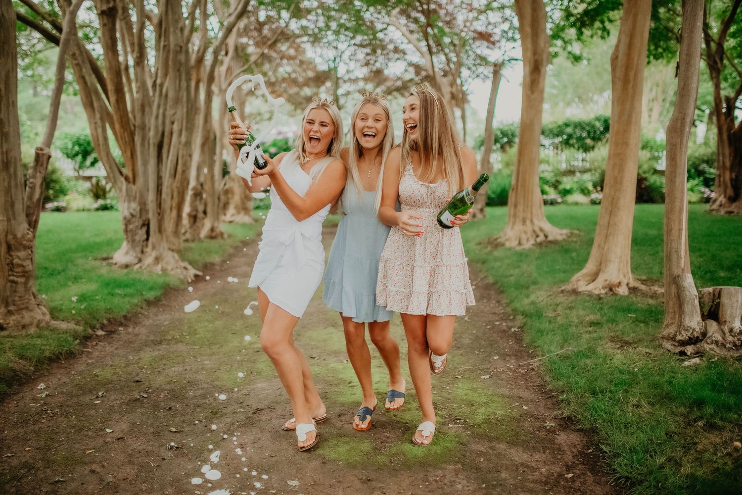Three young women laughing and popping champagne bottles on a tree-lined garden path.