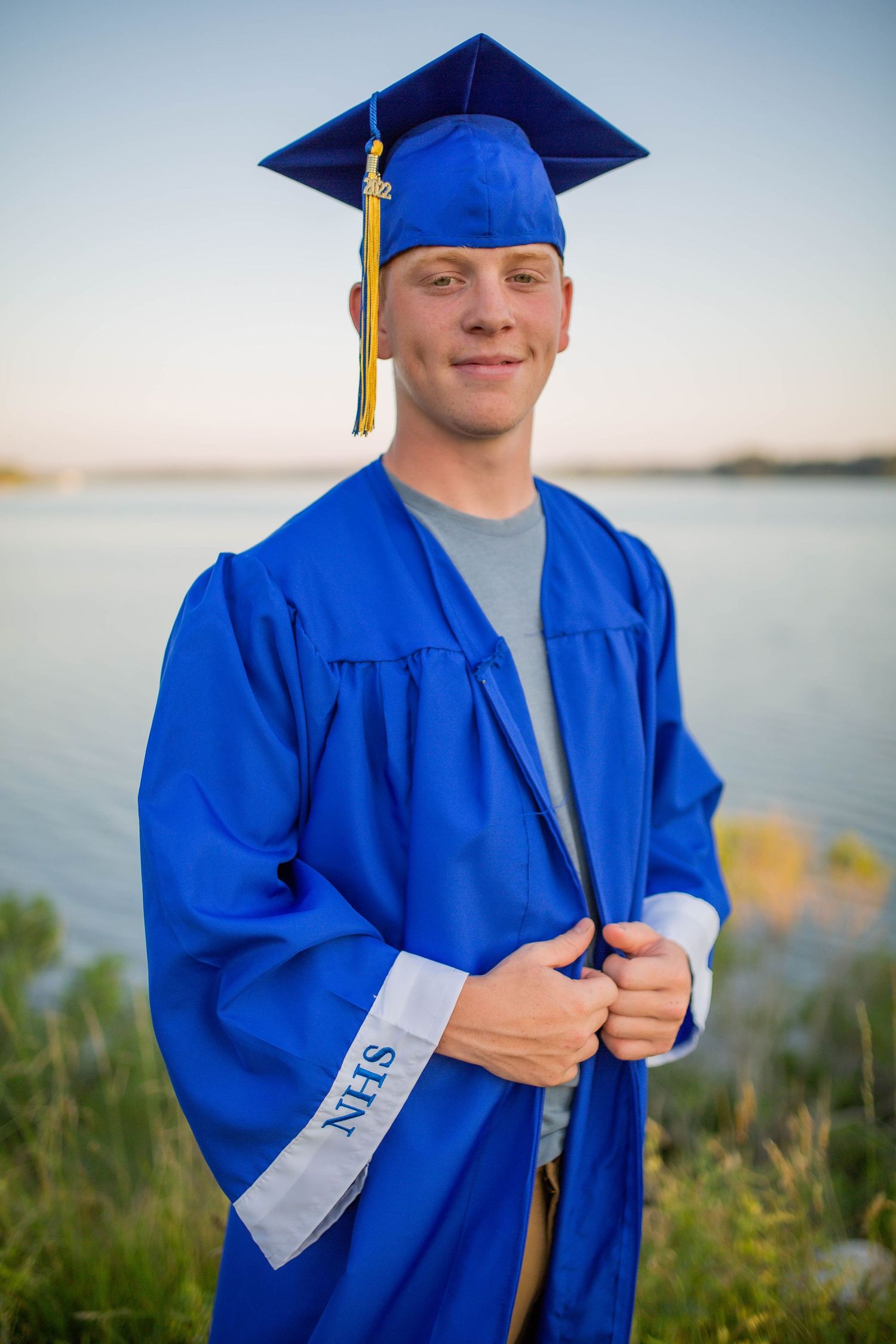 A smiling graduate in a royal blue cap and gown poses confidently by a lakeside at dusk.