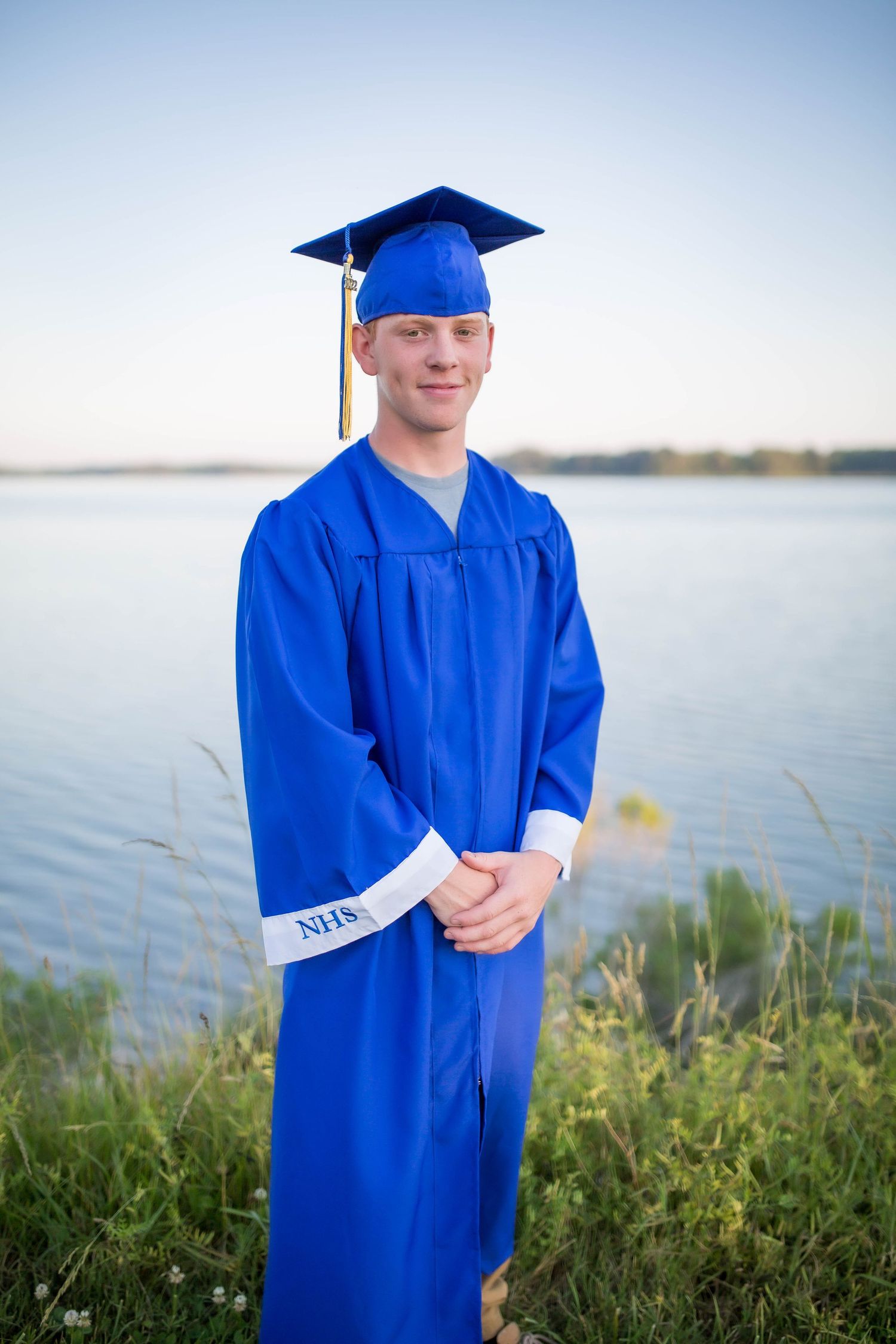 A young man in a royal blue graduation cap and gown stands near a calm lake at sunset.