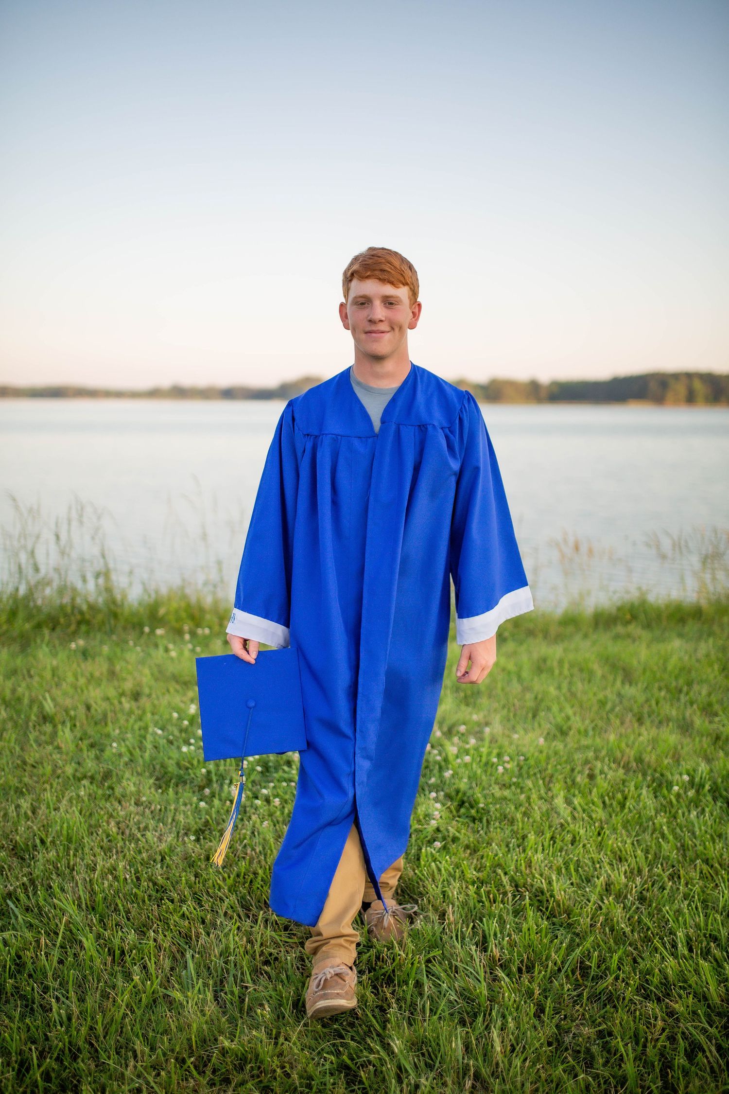 A red-haired young man in a blue graduation gown walks through green grass near a lake.