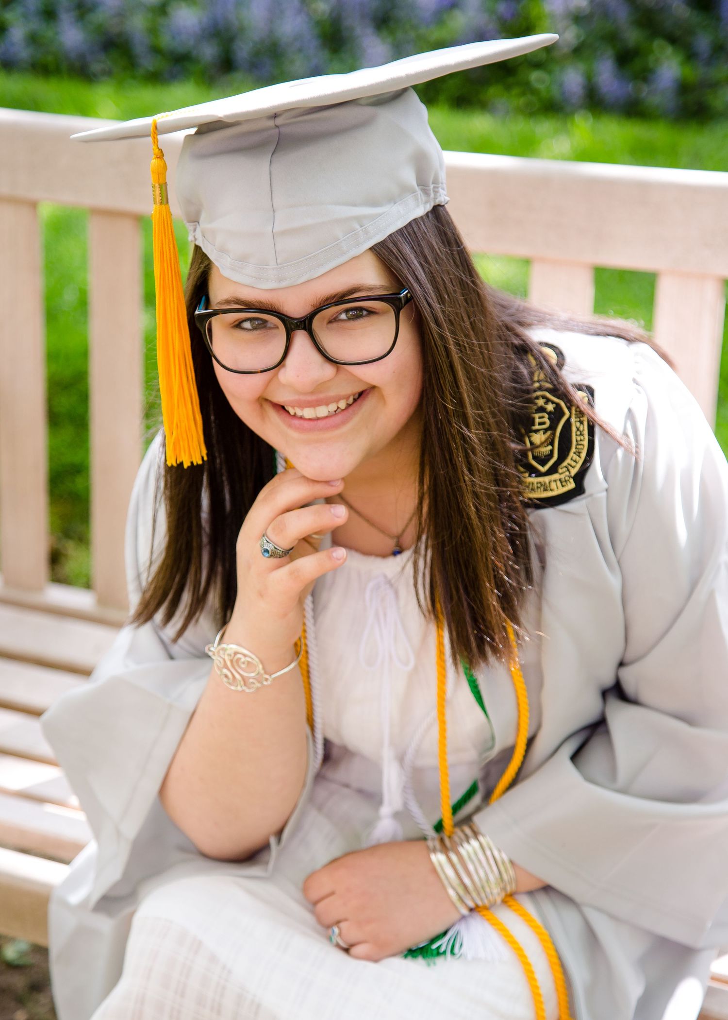 Smiling female graduate in gray cap and gown with yellow tassel sitting on bench.