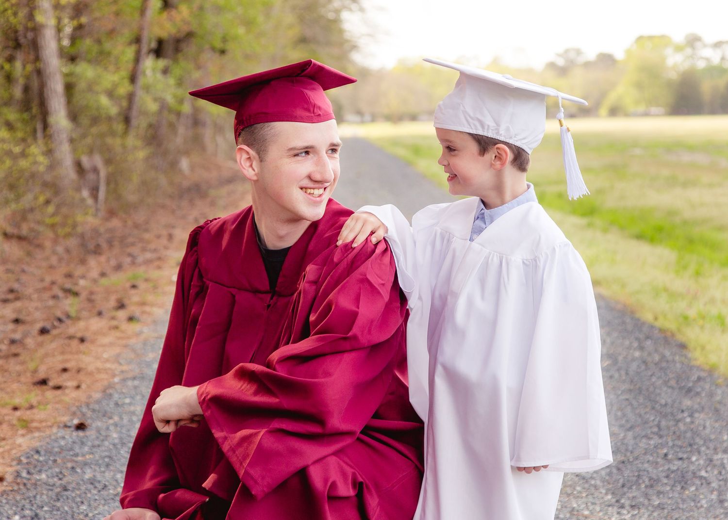 A high school graduate in burgundy cap and gown poses with a younger boy in white graduation attire.