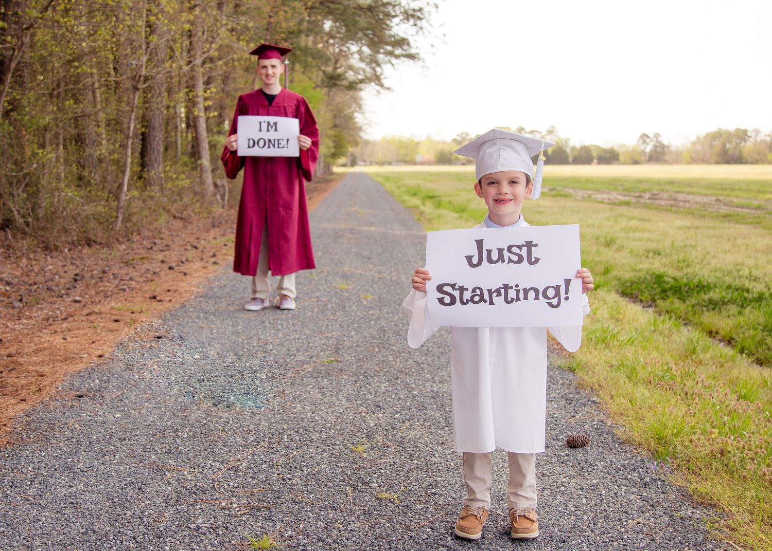Graduate holding I'm Done sign with young child holding Just Starting sign on rural path.