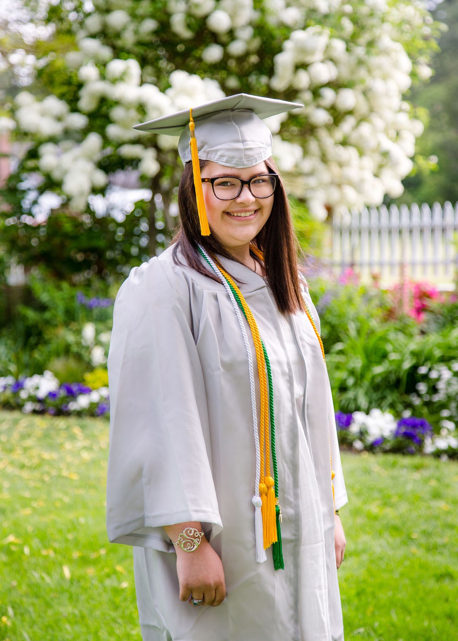 Female graduate in gray cap and gown with green and gold honor cords standing in garden.