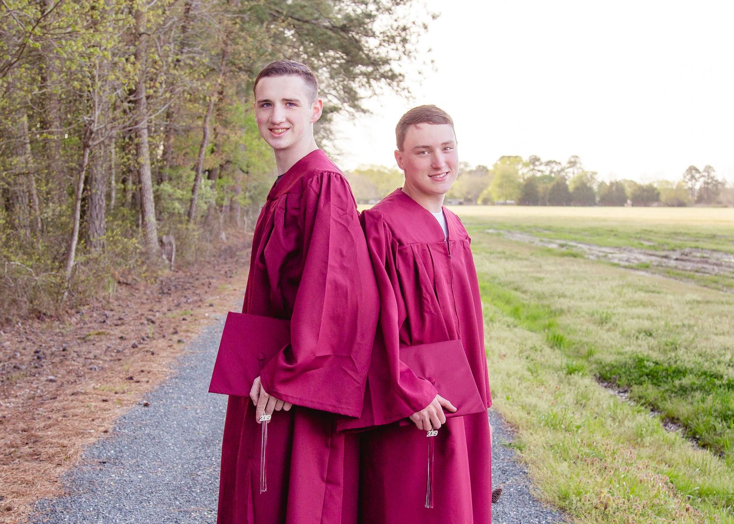Two young men in matching burgundy graduation gowns pose on a tree-lined gravel path.