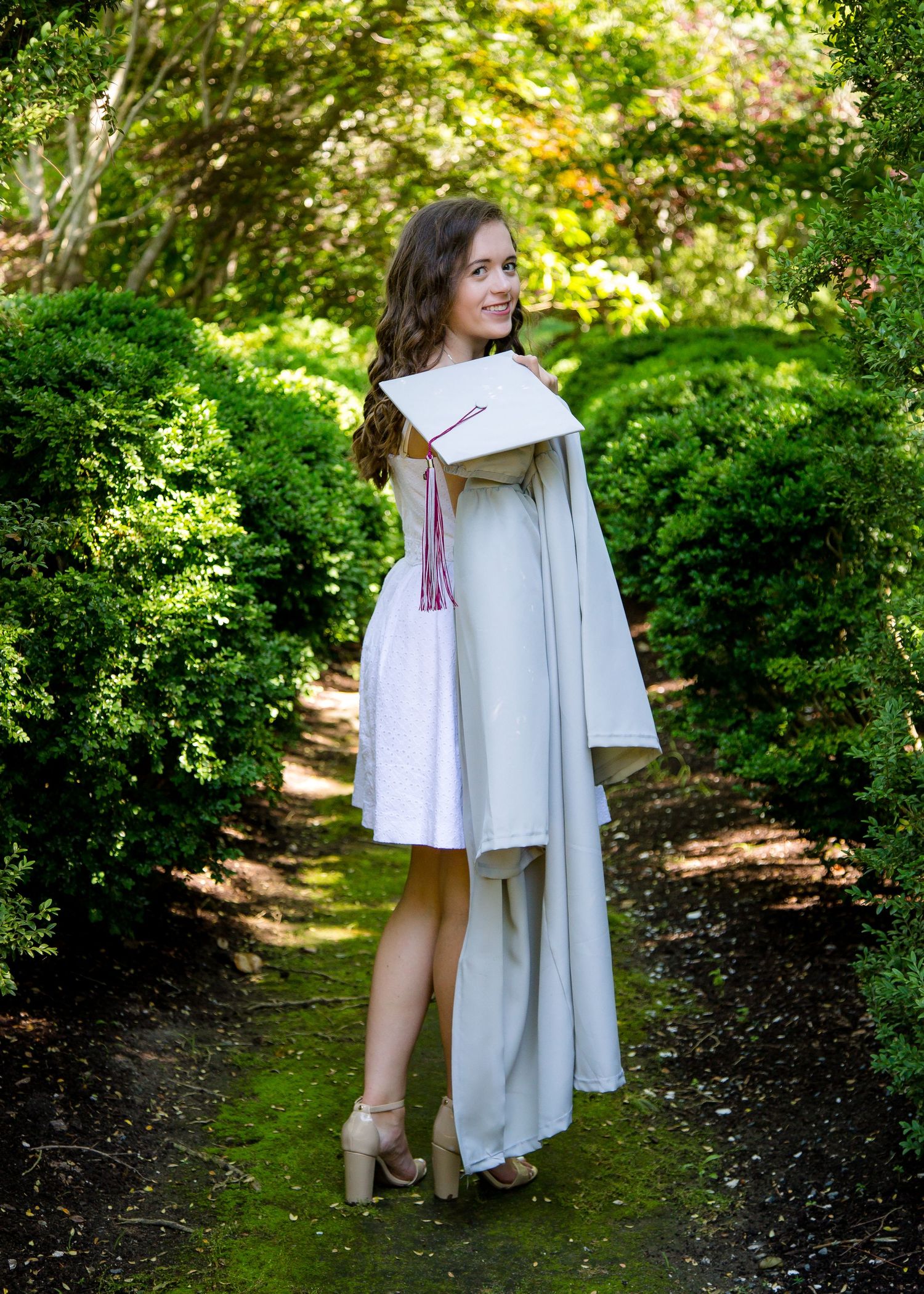 Female graduate in gray cap and gown walking through lush green garden path.