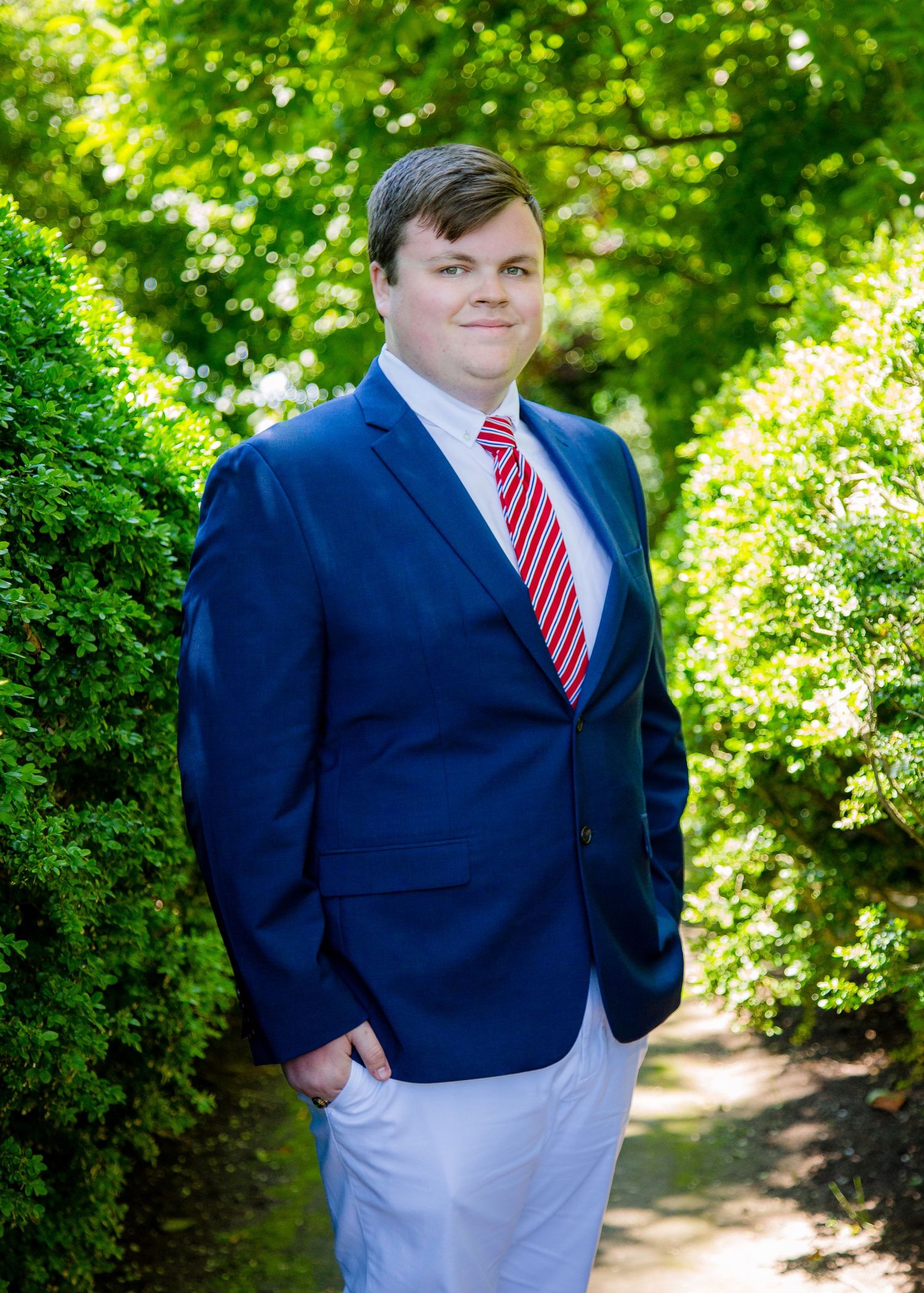 A young man in a navy blazer with red striped tie poses on a green tree-lined path.