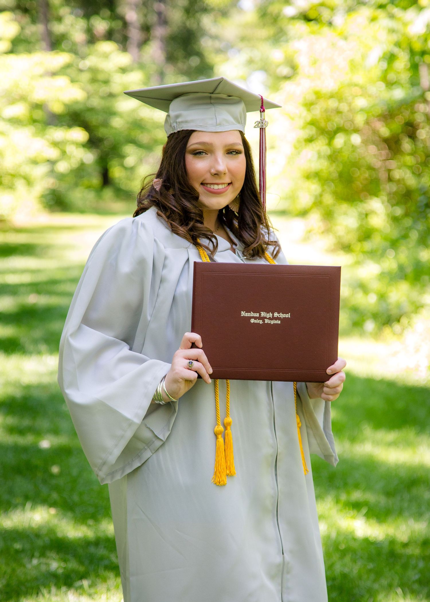 Female graduate in gray cap and gown holding burgundy diploma cover outdoors.