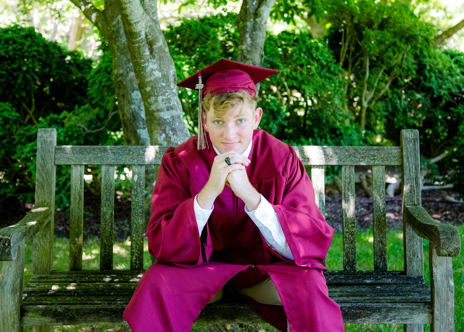 Male graduate in burgundy cap and gown sitting on wooden bench outdoors.